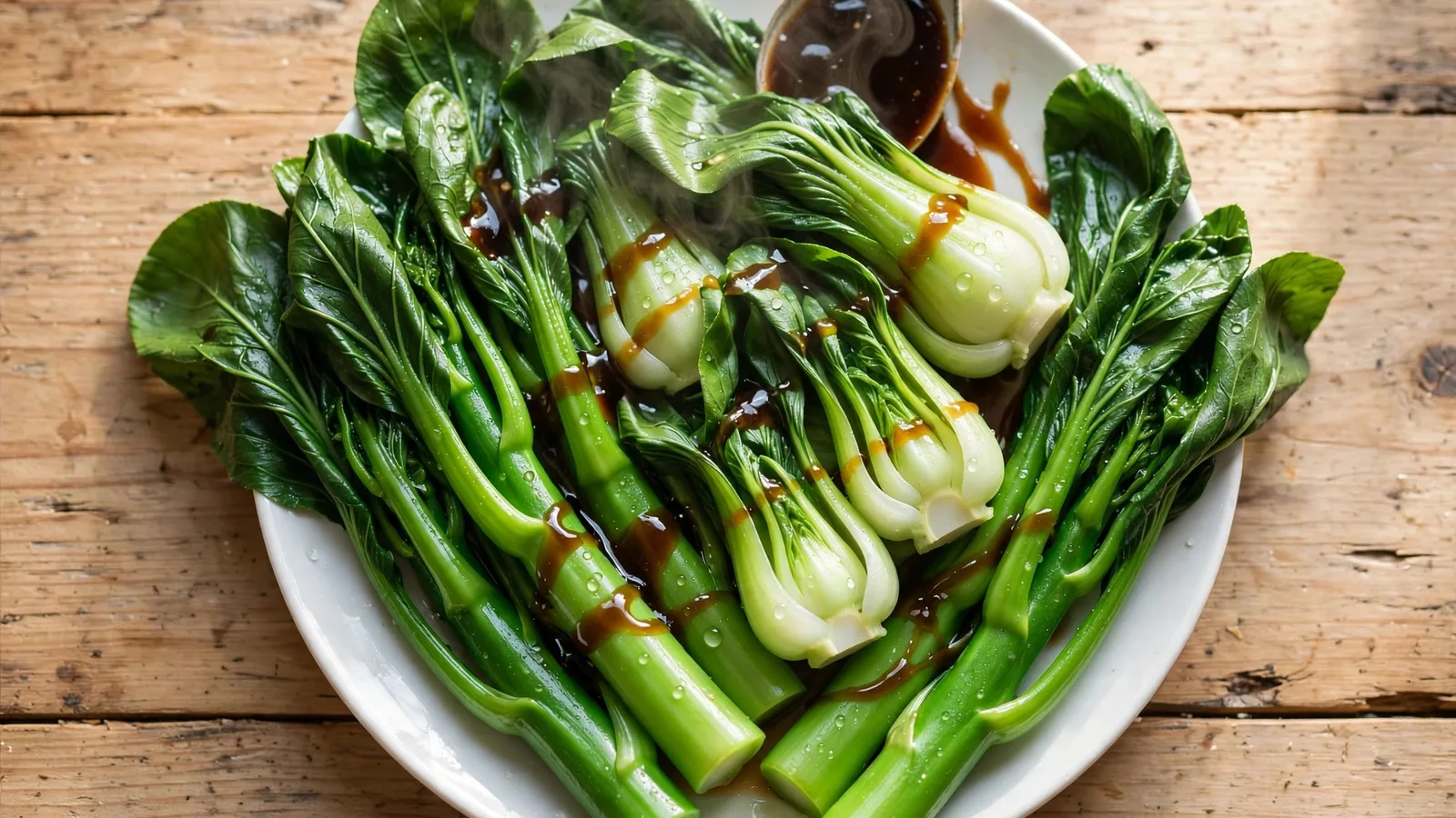 White plate of blanched gai lan and baby bok choy with vivid green stems and glossy leaves, dark oyster sauce being spooned over the top, steam rising, on a rustic wooden table