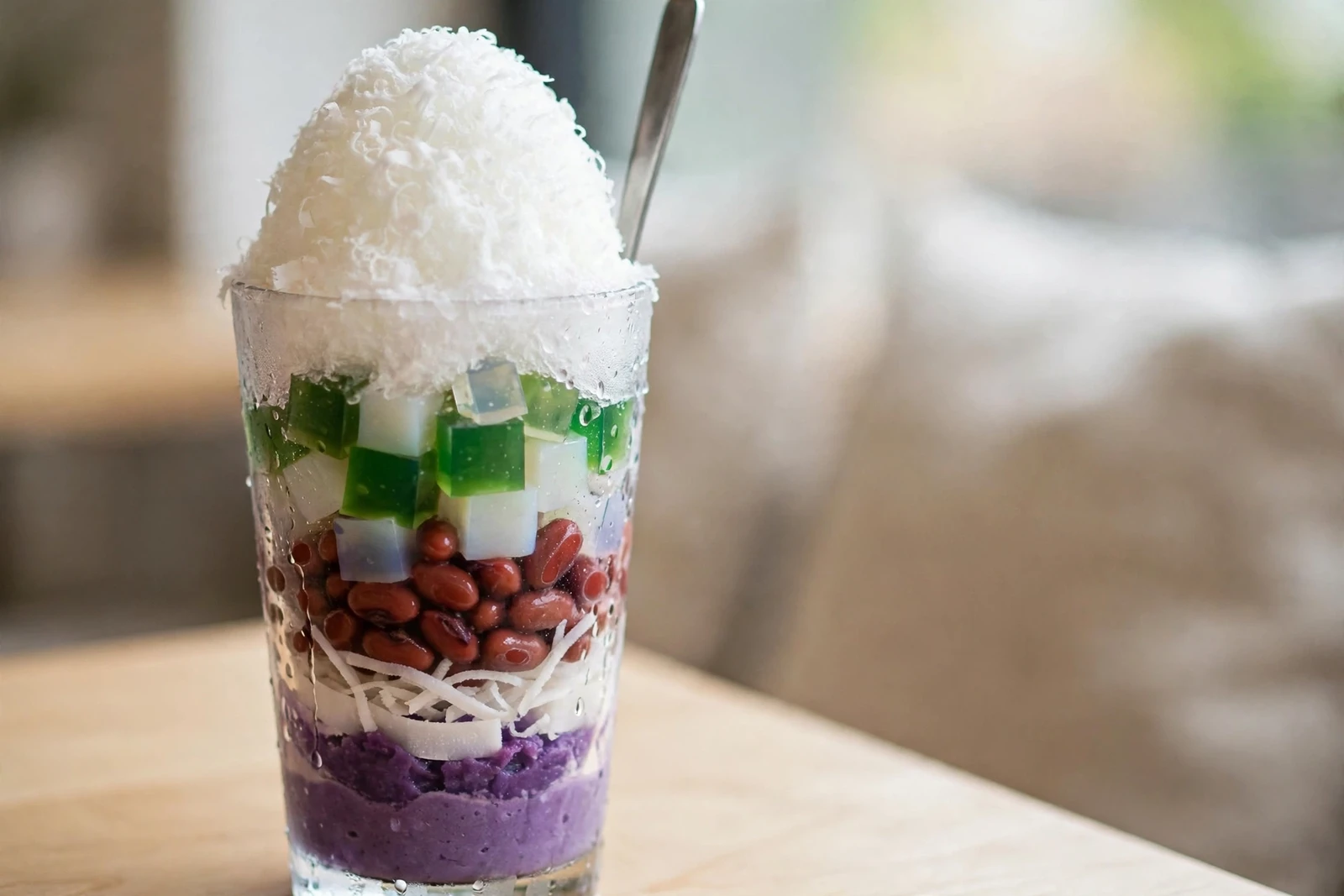 Side view of halo-halo layers in a tall glass: purple ube halaya at the bottom, white macapuno strips, red beans, green and white gelatin cubes, topped with a dome of shaved ice and a long metal spoon, condensation on the glass.