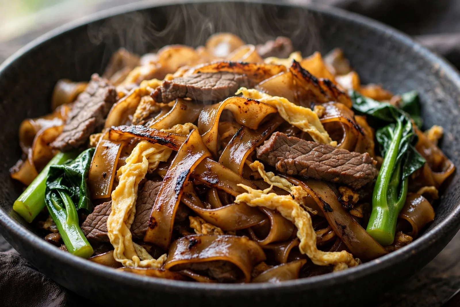 Close-up bowl of Pad See Ew with wide flat rice noodles caramelized in dark soy, beef slices, Chinese broccoli and scrambled egg